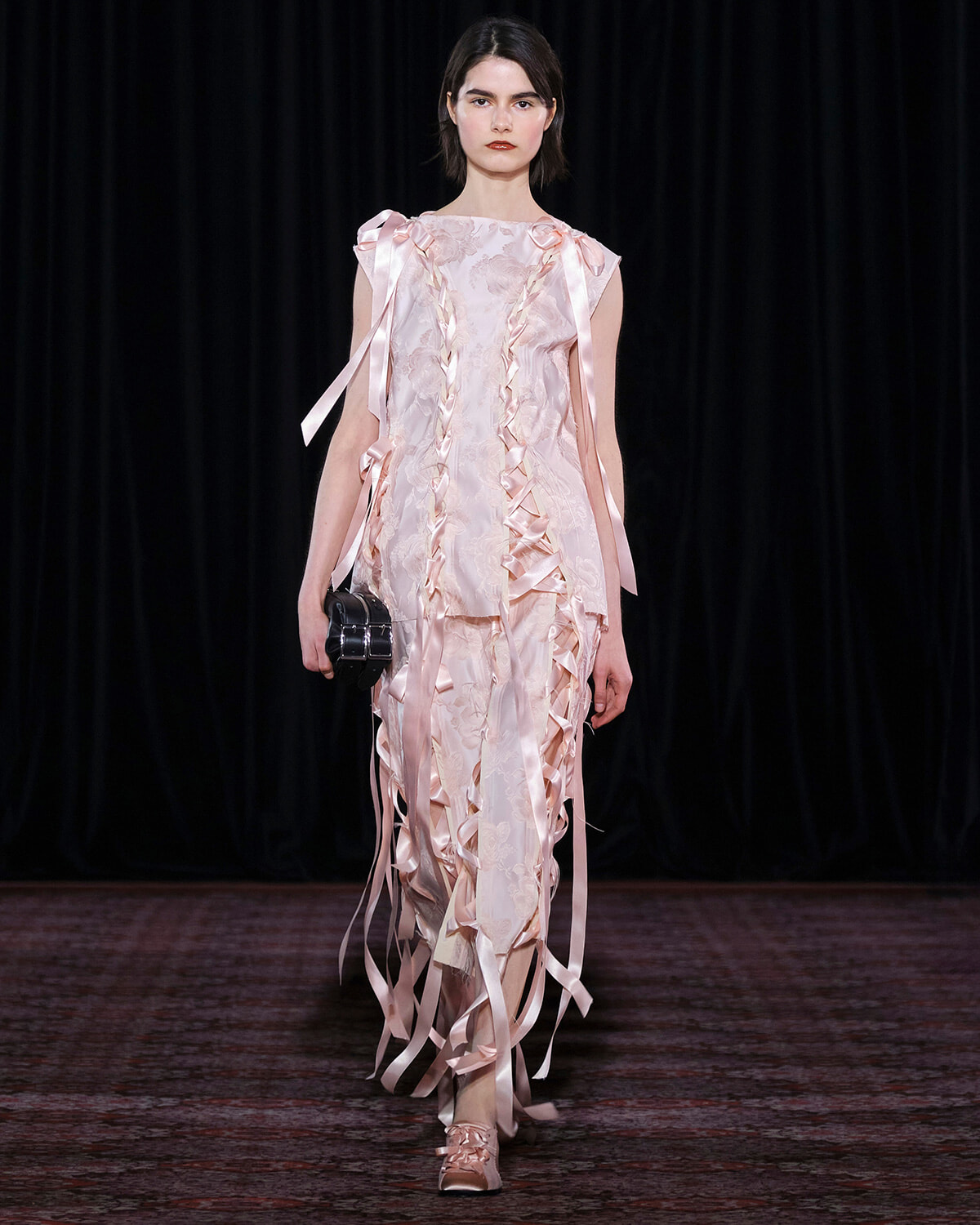 Female model walks the runway in a pale pink, ruffled gown with satin ribbons and a small black clutch.