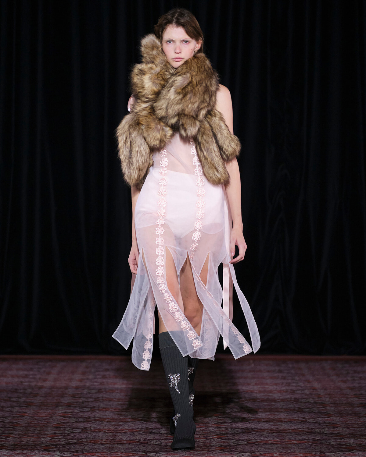 Model on a runway wearing a large fur stole over a sheer pink gown with lace panels and embellished socks.