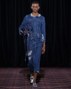 Female model walks the runway in a dark-blue denim suit with a distressed jeans and a jeweled collar accessory.