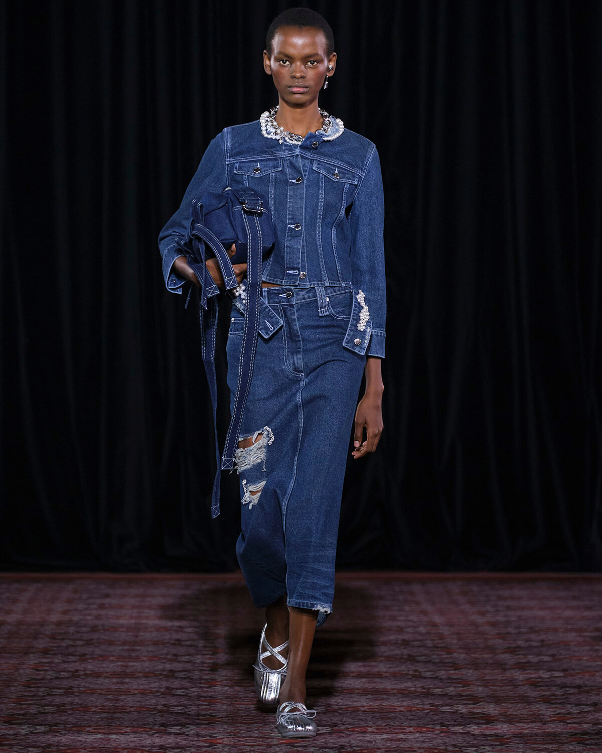 Female model walks the runway in a dark-blue denim suit with a distressed jeans and a jeweled collar accessory.