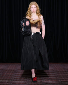 Model on a runway wearing a fur bandeau top, black wide-leg pants, a chain belt, and a black sculptural handbag.