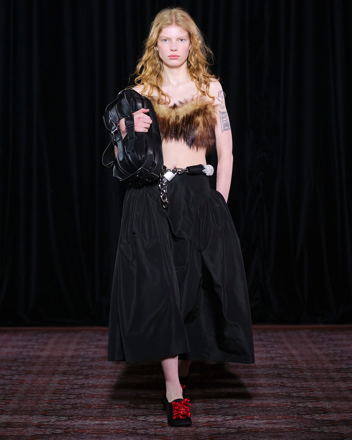 Model on a runway wearing a fur bandeau top, black wide-leg pants, a chain belt, and a black sculptural handbag.
