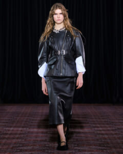 Model walking a runway in a black leather belted jacket with oversized white cuff sleeves and a black midi skirt, dark backdrop behind.