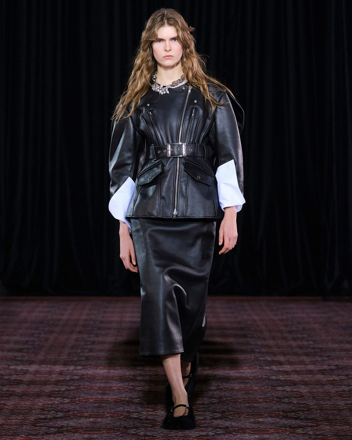 Model walking a runway in a black leather belted jacket with oversized white cuff sleeves and a black midi skirt, dark backdrop behind.