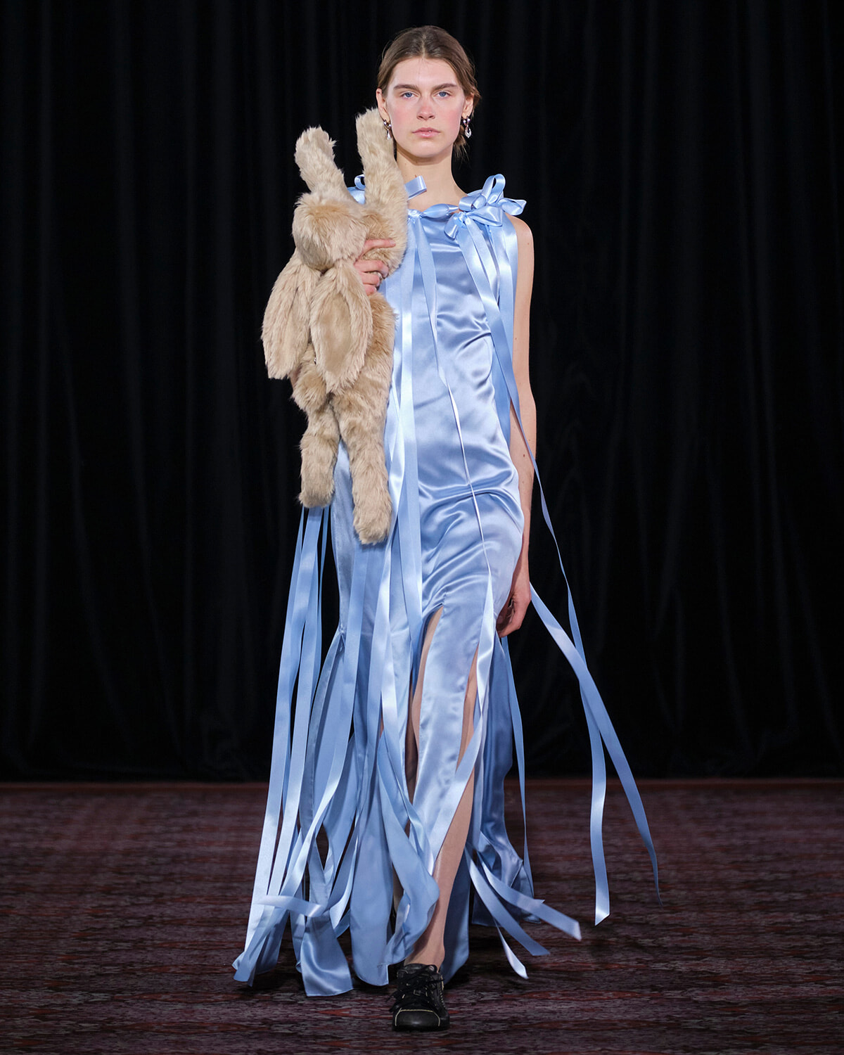 Model on a runway wearing a light blue satin gown with ribbon fringe, holding a beige plush bunny.