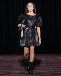 Model walking a fashion show wearing a black leather dress with puff sleeves, belt, and pearl handbag against a dark backdrop.