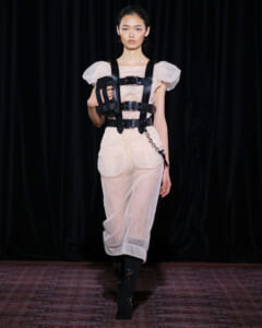 Runway model wearing a sheer beige jumpsuit with a black leather harness and matching arm pouch, walking forward with a neutral expression.