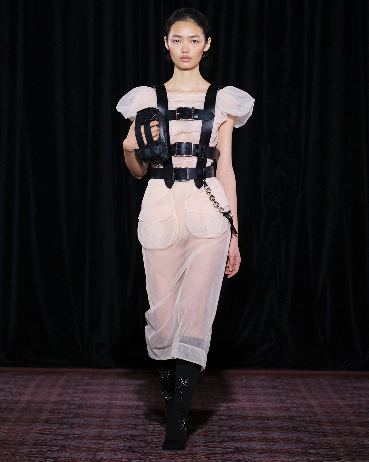 Runway model wearing a sheer beige jumpsuit with a black leather harness and matching arm pouch, walking forward with a neutral expression.