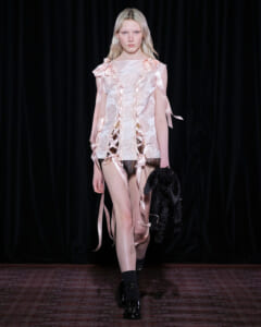 Model walks a runway in a pale pink lace dress with satin ribbon ties, carrying a black jacket in her left hand.