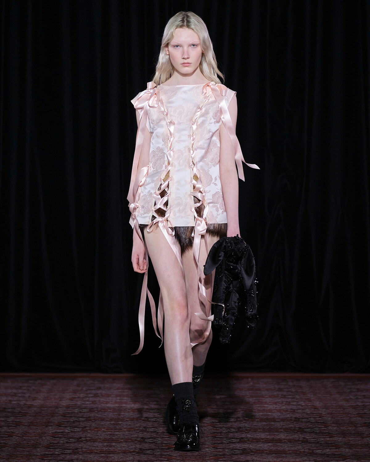 Model walks a runway in a pale pink lace dress with satin ribbon ties, carrying a black jacket in her left hand.