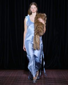 Model in a light blue satin jumpsuit walking the runway, draped with a brown fur stole over one shoulder.