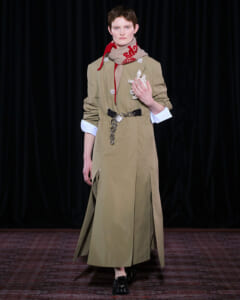 Model on a runway wearing a knee-length tan trench dress with rolled sleeves, a red scarf, and a chain belt.