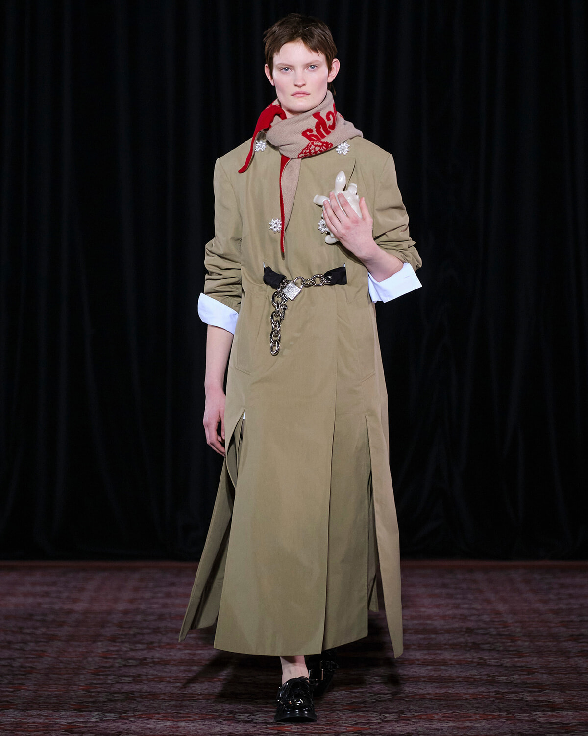 Model on a runway wearing a knee-length tan trench dress with rolled sleeves, a red scarf, and a chain belt.