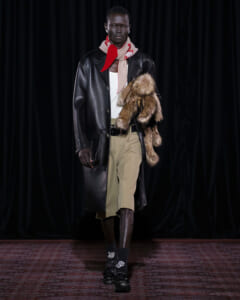 Male model on a runway wearing a long black leather coat, white top, tan cropped trousers, and a red scarf with a fur stole draped over the shoulder.