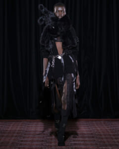 Model on a runway wearing an avant-garde black ensemble with a voluminous fur neckpiece and a chained belt.