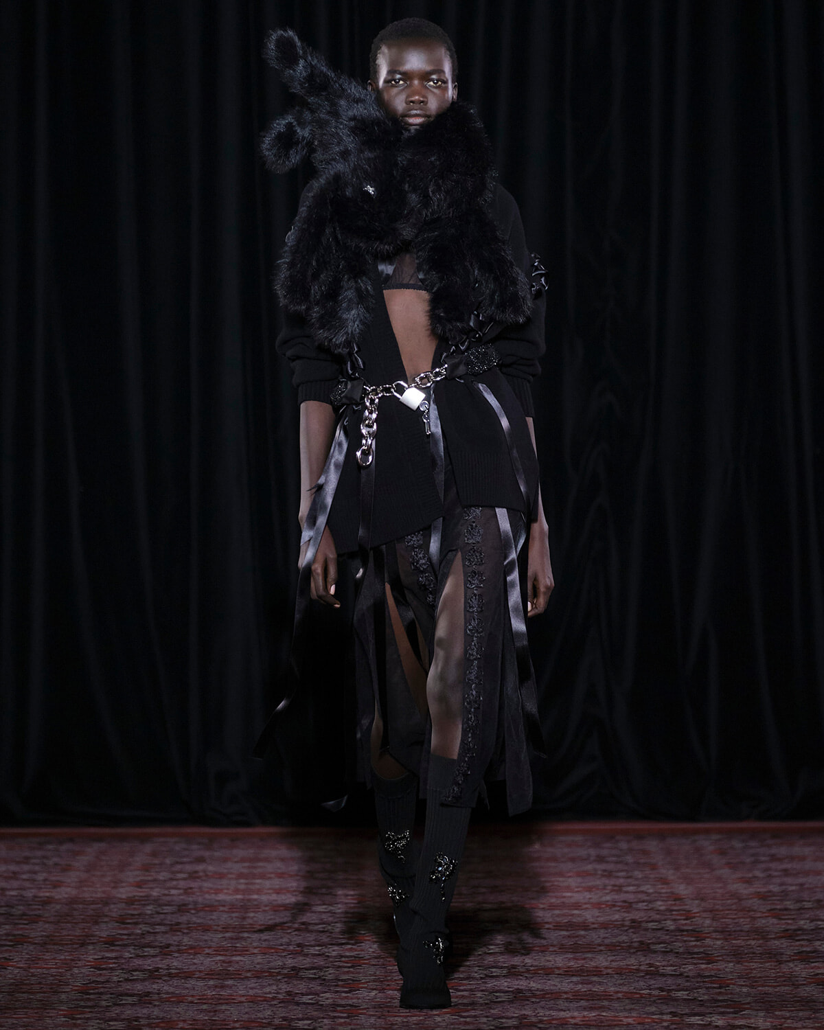 Model on a runway wearing an avant-garde black ensemble with a voluminous fur neckpiece and a chained belt.