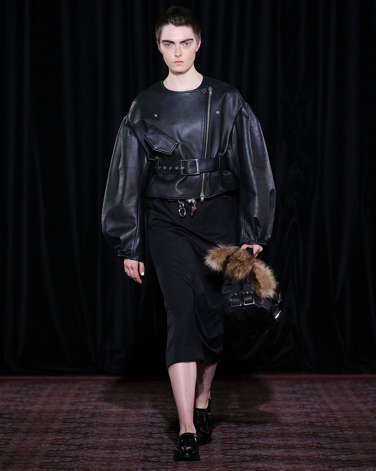 Model walks the runway in an oversized black leather jacket with a waist belt, paired with black cropped pants and a fur-trimmed accessory held in one hand