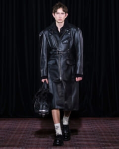 Male model wearing a long black leather trench coat with a belted waist, carrying a black fur-trim bag on a runway.