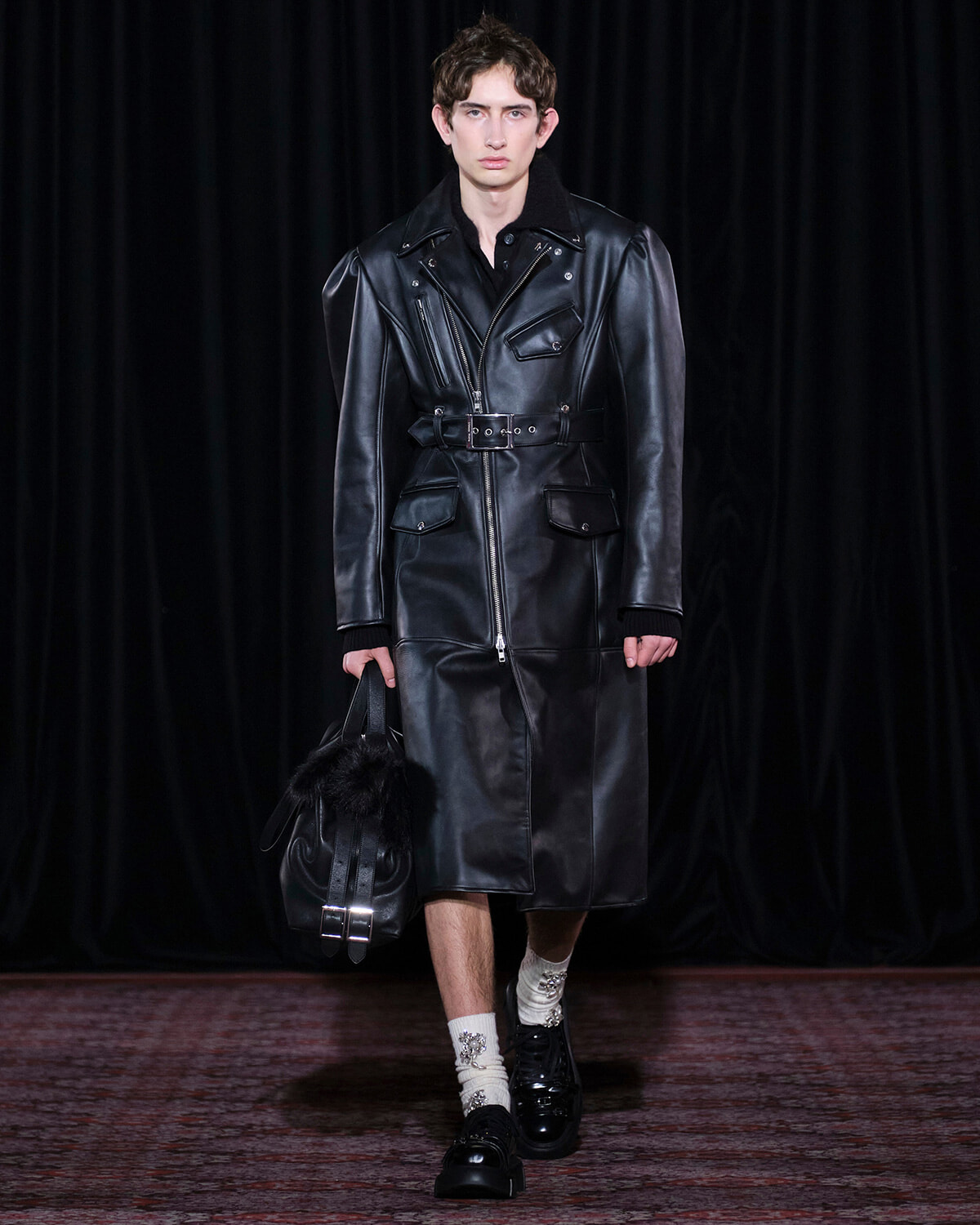 Male model wearing a long black leather trench coat with a belted waist, carrying a black fur-trim bag on a runway.