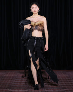 Model walking a runway in a black fringe skirt with a fur bandeau top, holding a black coat over her arm.