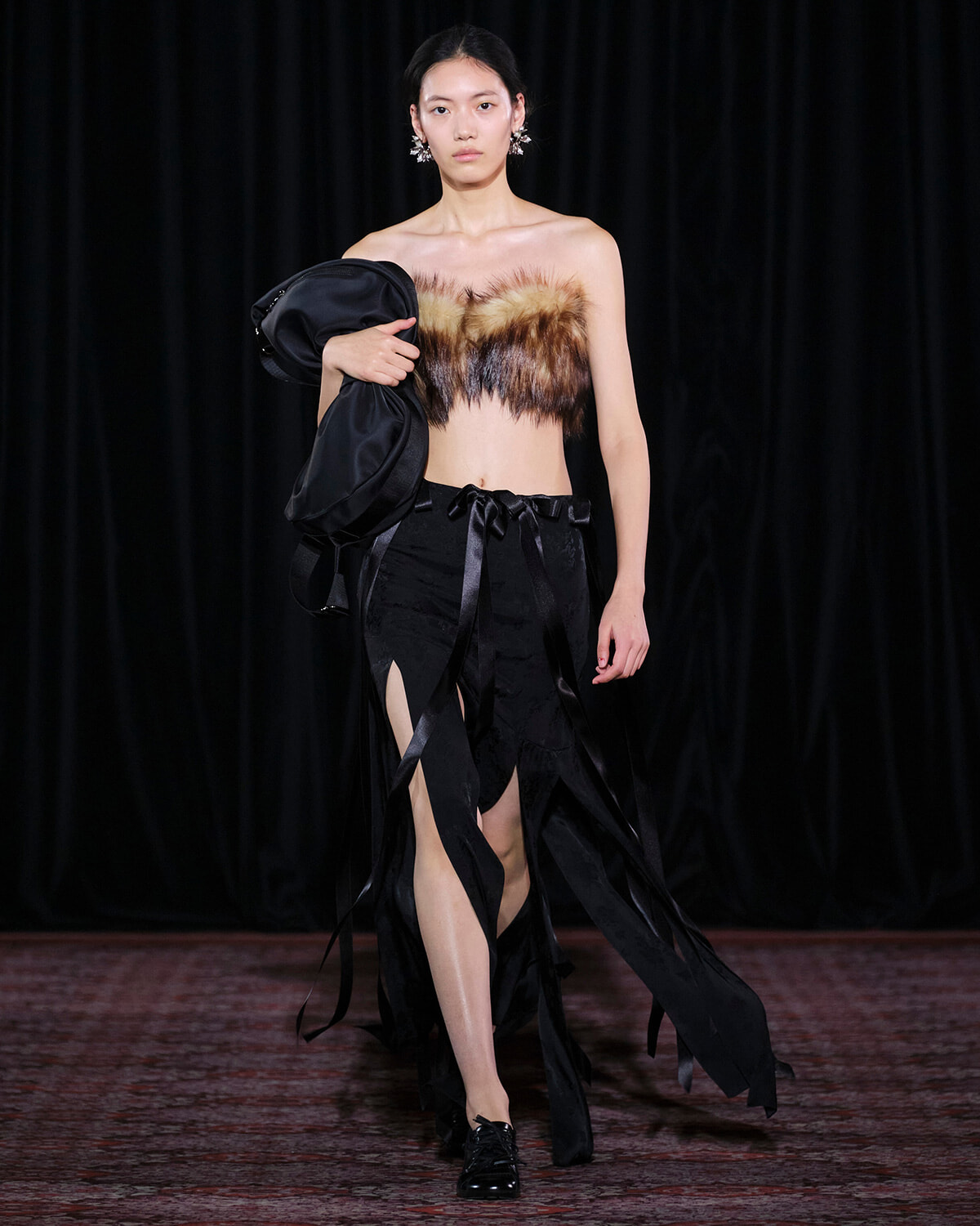 Model walking a runway in a black fringe skirt with a fur bandeau top, holding a black coat over her arm.