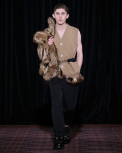 Male model walking a runway in a sleeveless beige knit top, fur stole draped over his shoulder, and black trousers with a belt and boots.