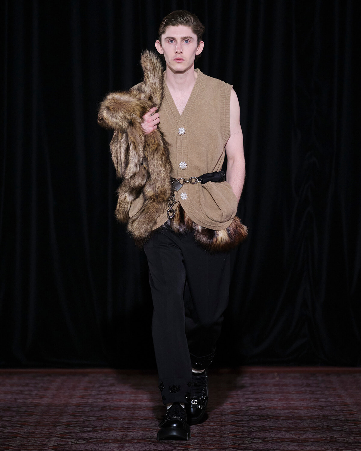 Male model walking a runway in a sleeveless beige knit top, fur stole draped over his shoulder, and black trousers with a belt and boots.