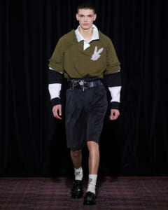 Male model on a runway wearing an olive polo with ruffle details and a white collar, paired with black belted knee-length shorts and white accented socks with black loafers.