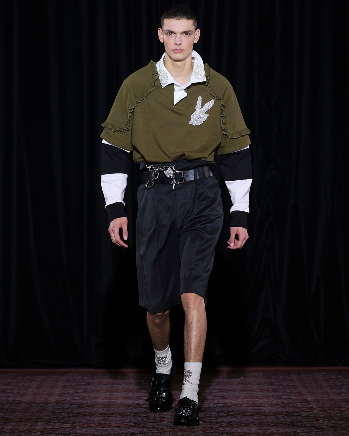 Male model on a runway wearing an olive polo with ruffle details and a white collar, paired with black belted knee-length shorts and white accented socks with black loafers.