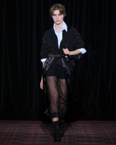 Model on a runway wearing an oversized black cardigan over a white shirt, with a sheer black maxi skirt and chain-adorned belt.
