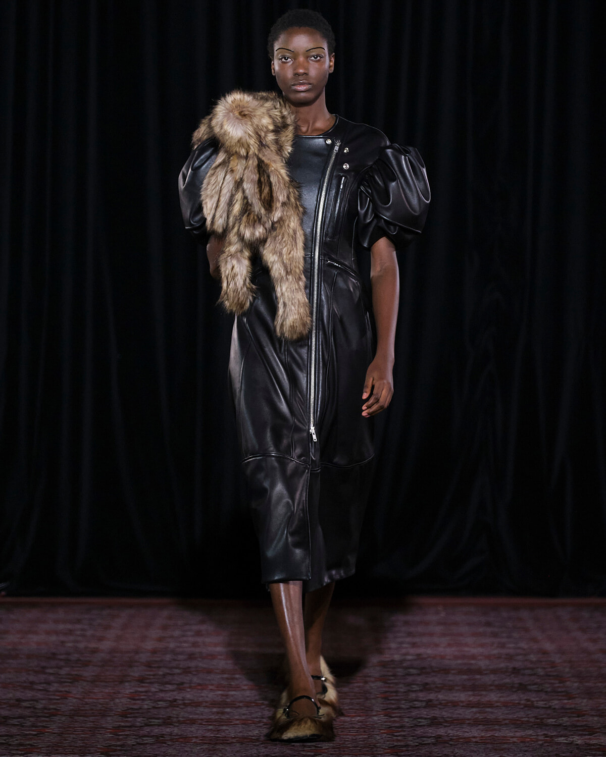 Model walks a runway in a black leather dress with puffed sleeves, holding a fur stole over her shoulder against a dark backdrop.