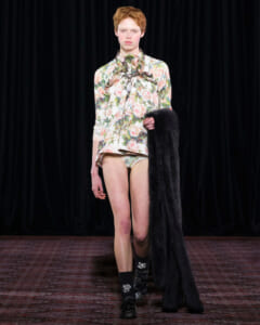 Model walking a runway in a floral print blouse and matching shorts, holding a long black fur coat over one arm.