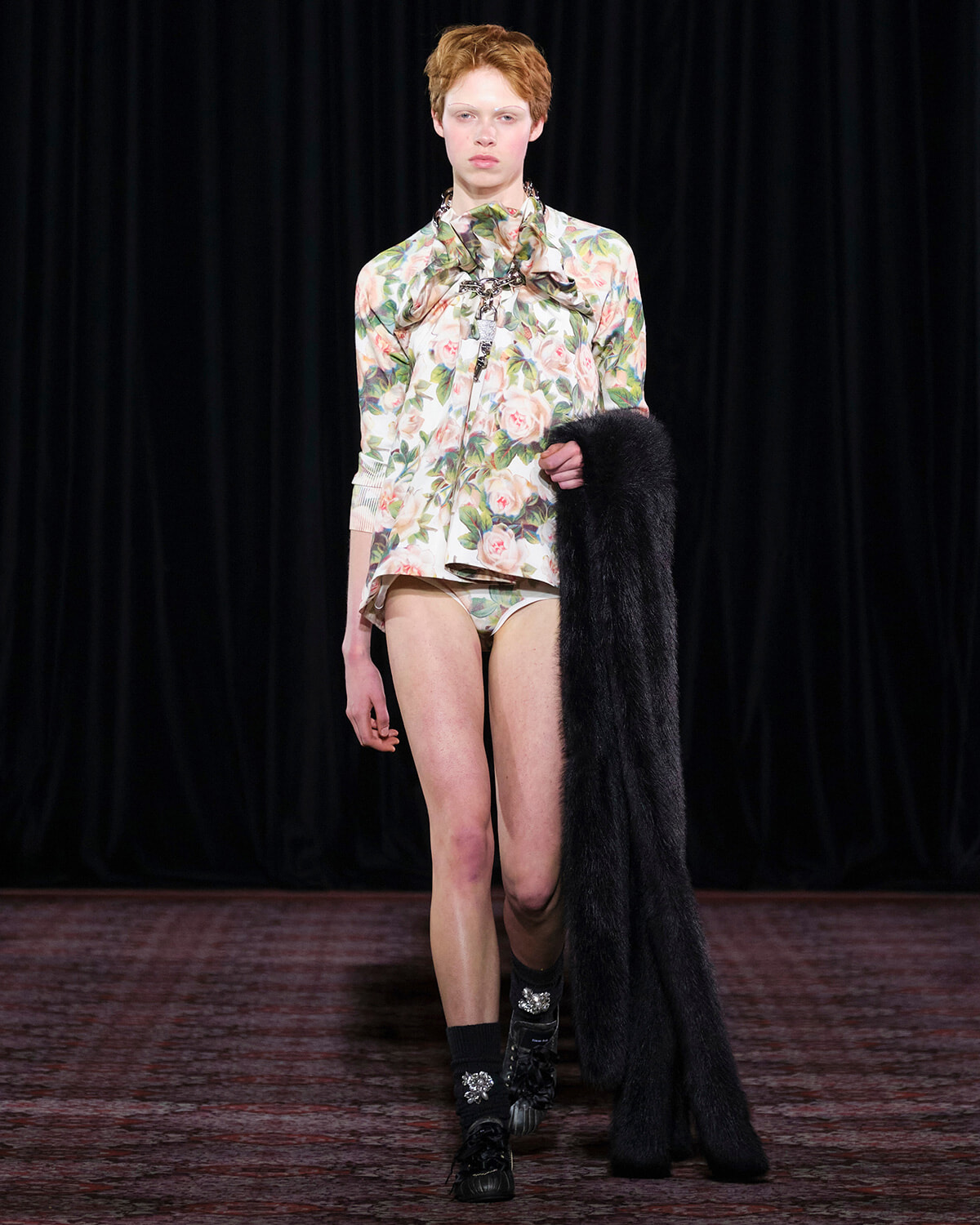 Model walking a runway in a floral print blouse and matching shorts, holding a long black fur coat over one arm.