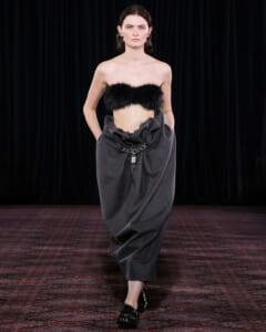 Model walking a runway in a strapless black fur bandeau and a voluminous, draped charcoal gray skirt with a metal chain belt.