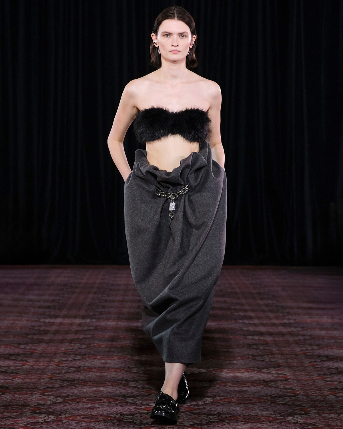 Model walking a runway in a strapless black fur bandeau and a voluminous, draped charcoal gray skirt with a metal chain belt.