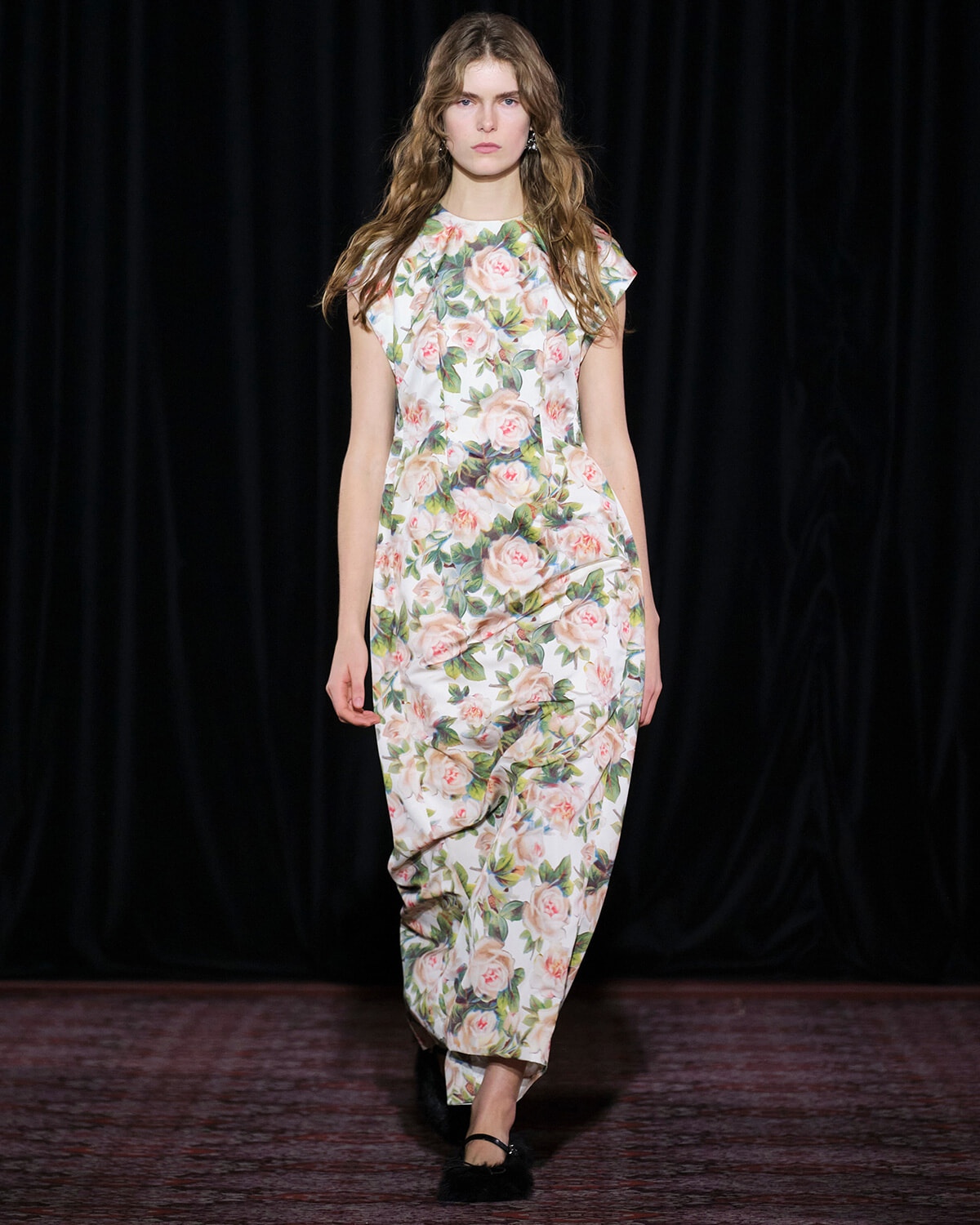 Model walking a pale floral print dress with pink roses down a dark runway backdrop.