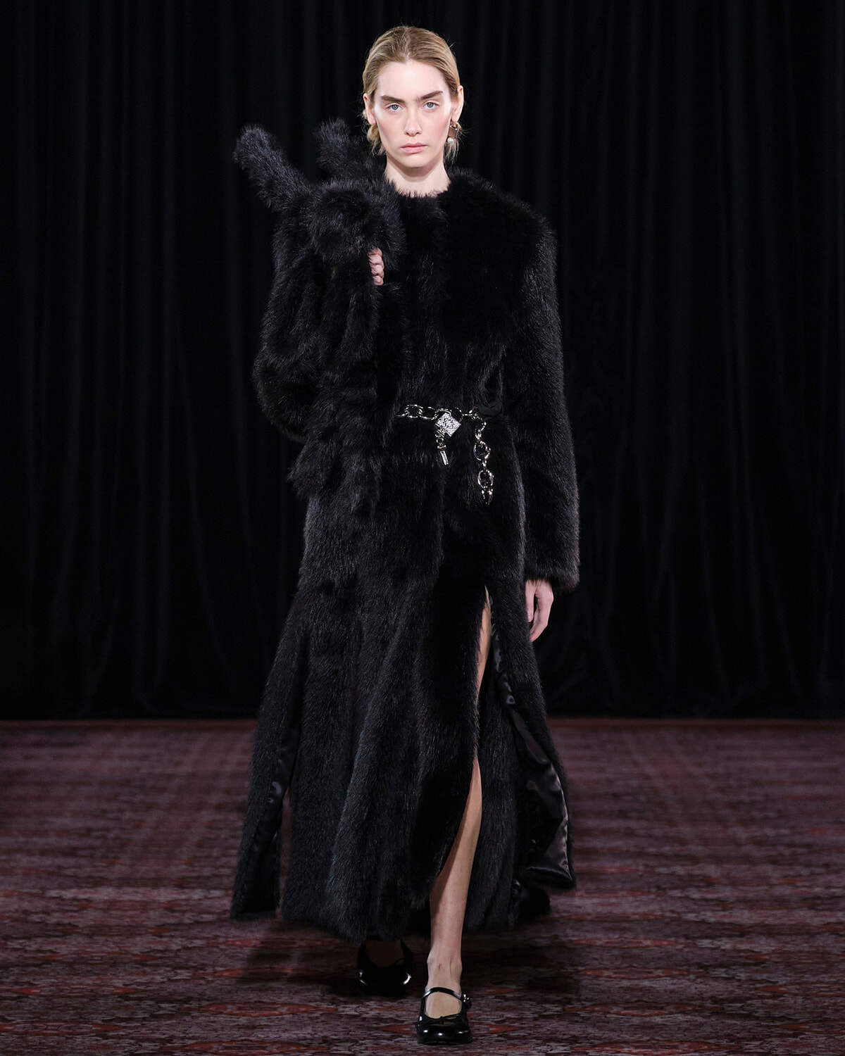 Model walks the runway in a long black fur coat cinched with a silver belt, with a high slit showing her leg.