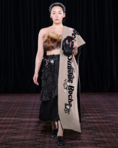 Model on a runway wearing a fur bandeau top, sparkling black skirt, a beige scarf draped over one shoulder, and a small black handbag.