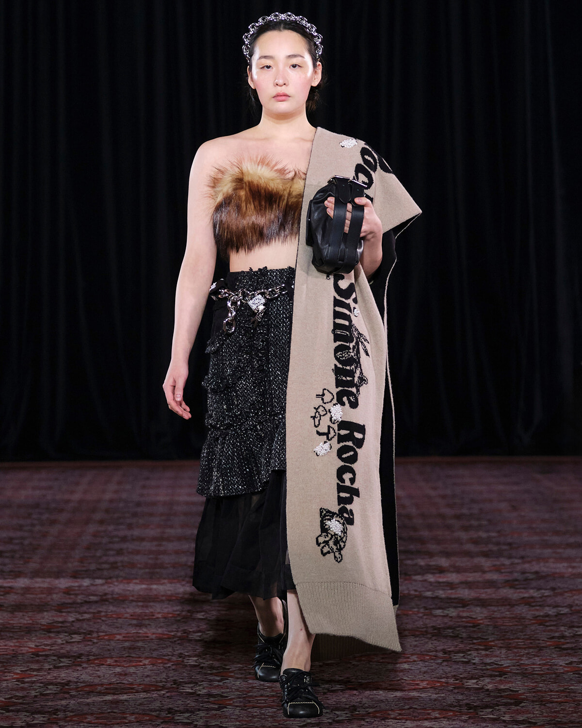 Model on a runway wearing a fur bandeau top, sparkling black skirt, a beige scarf draped over one shoulder, and a small black handbag.