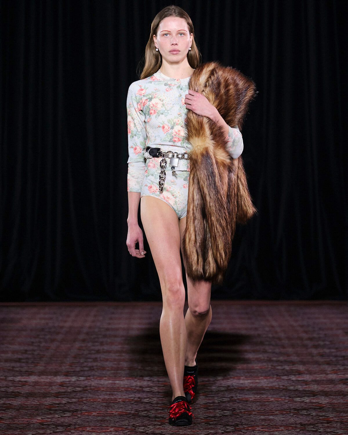 Model walking a runway in a pastel floral bodysuit, cinched with a chunky belt, while holding a brown fur stole over her shoulder.