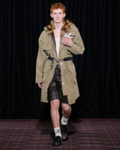 Male model on a fashion runway wearing a tan parka with fur-trimmed hood, belt, and brown leather shorts; holding a small gray plush rabbit.