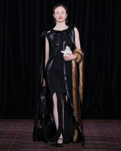 Woman in a black, ribbon-accented gown on a stage, holding a white sculpture and a fur stole draped over her arm.