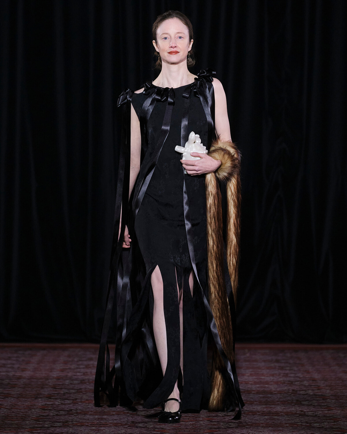 Woman in a black, ribbon-accented gown on a stage, holding a white sculpture and a fur stole draped over her arm.