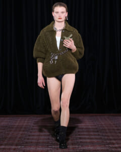 Model walking a runway in an oversized olive knit cardigan with a chain belt, black socks, and patent shoes, holding a small sculpture against a dark backdrop.