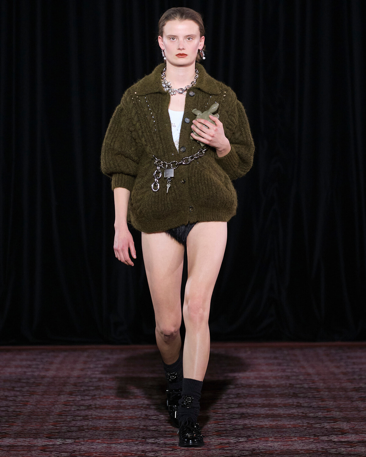 Model walking a runway in an oversized olive knit cardigan with a chain belt, black socks, and patent shoes, holding a small sculpture against a dark backdrop.