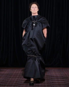 Model walks a runway in a dramatic black oversized dress with voluminous shoulders and gathered waist featuring chain jewelry.