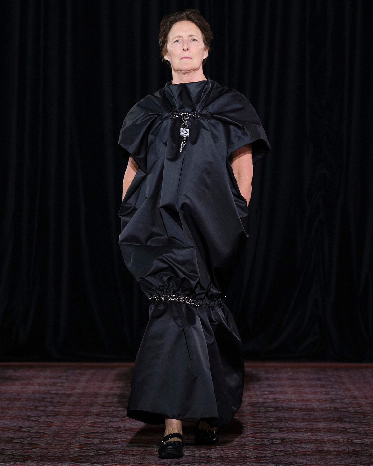 Model walks a runway in a dramatic black oversized dress with voluminous shoulders and gathered waist featuring chain jewelry.