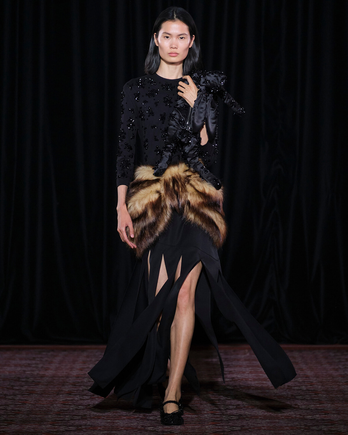 Model walks the runway in a black, beaded top paired with a fur belt and a long black skirt with dramatic slits on both sides.