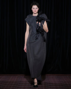 Model walks the runway in a long gray dress with structured shoulders, holding a large black sculptural ornament.