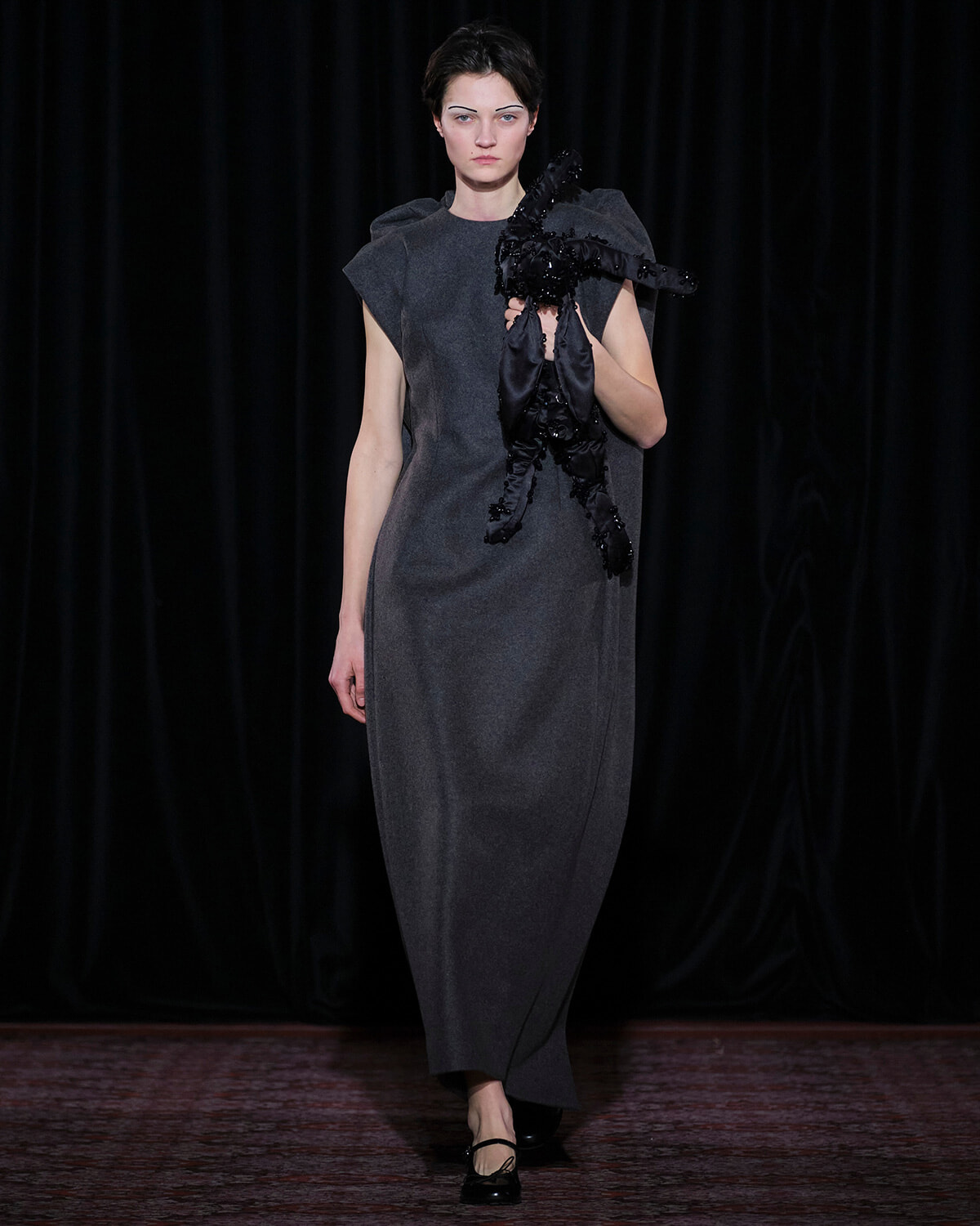 Model walks the runway in a long gray dress with structured shoulders, holding a large black sculptural ornament.